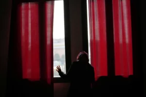 Woman looking out a window. Long red curtains hang on either side. 